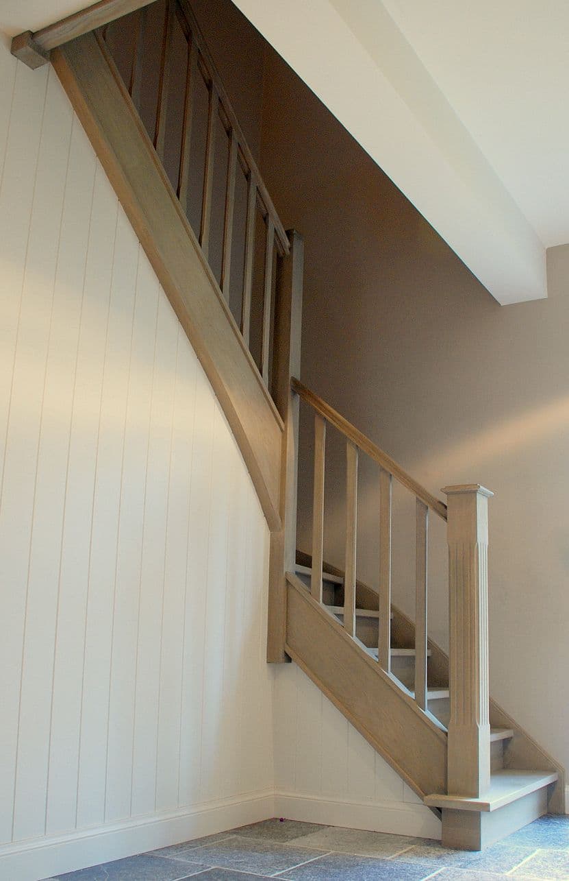 Houten trap met leuning en spijlen, witte wandpanelen links, donkere muur en stenen vloer.