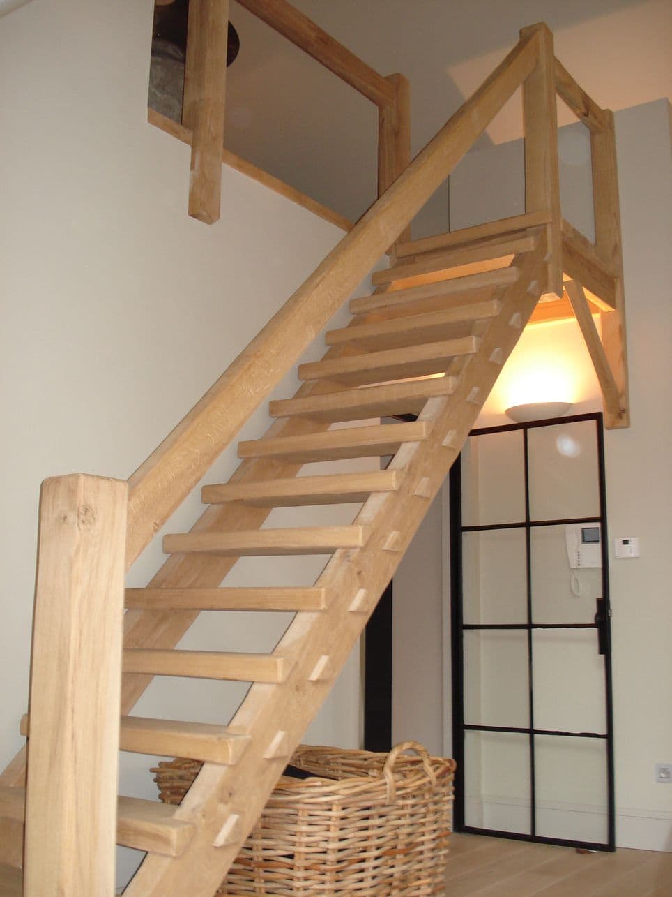Houten trap met open treden naar bovenverdieping, naast een glazen deur met zwart frame; vooraan een rieten mand.