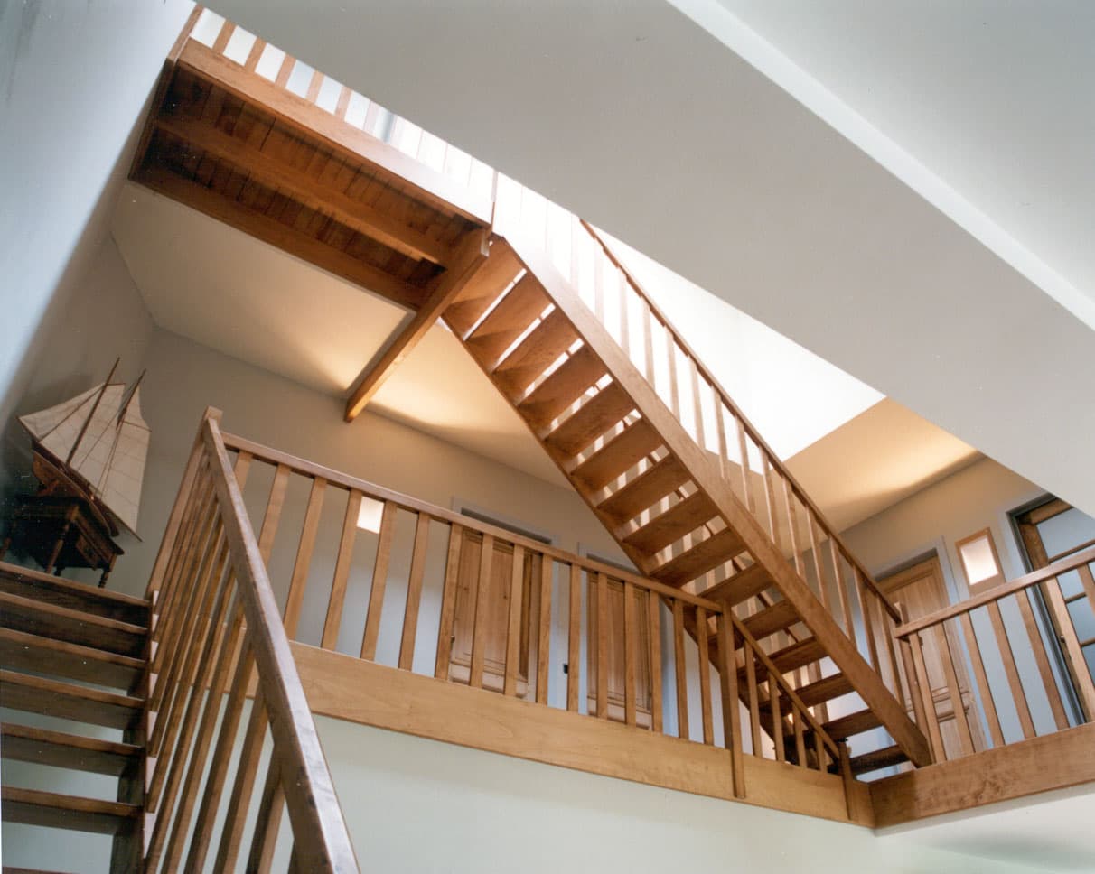Interieur met houten trap en balkon met leuningen, meerdere verdiepingen en veel licht; links staat een modeljacht op een plank.
