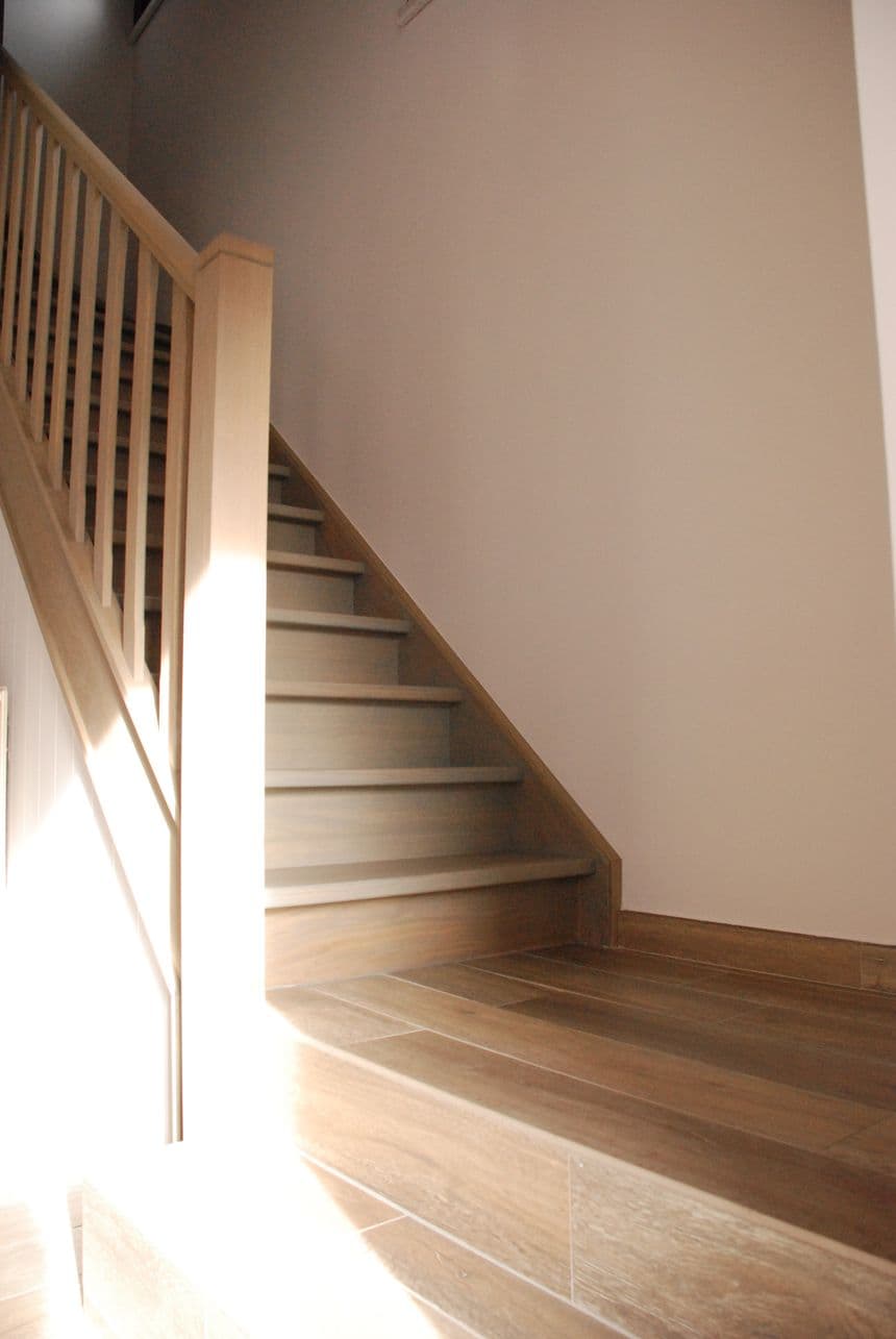 Houten trap met leuning links, beige muur rechts, houten vloer en zonlicht op de vloer.