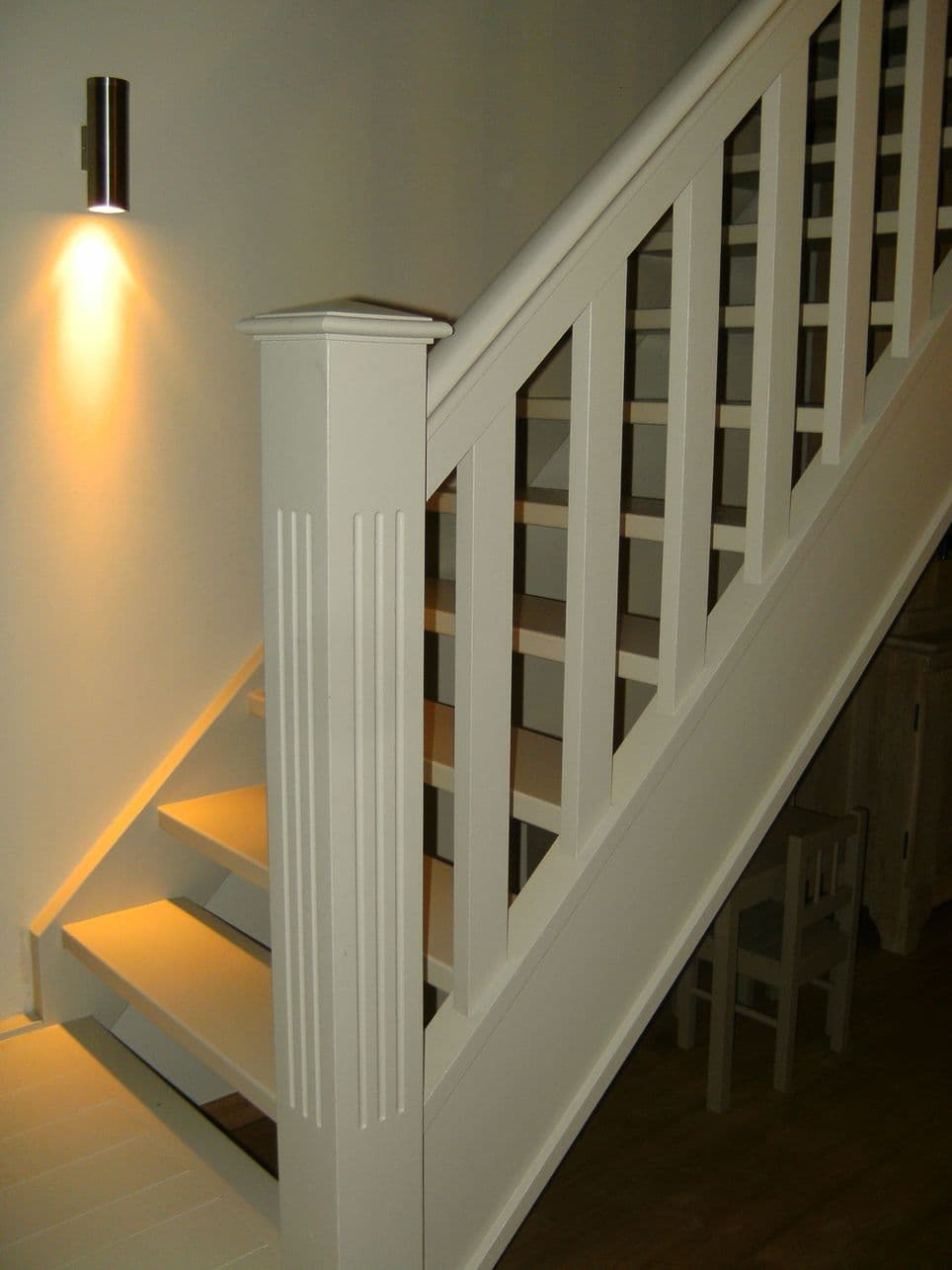 Witte trapleuning met verticale spijlen langs een binnentrap, met muurlicht erboven.