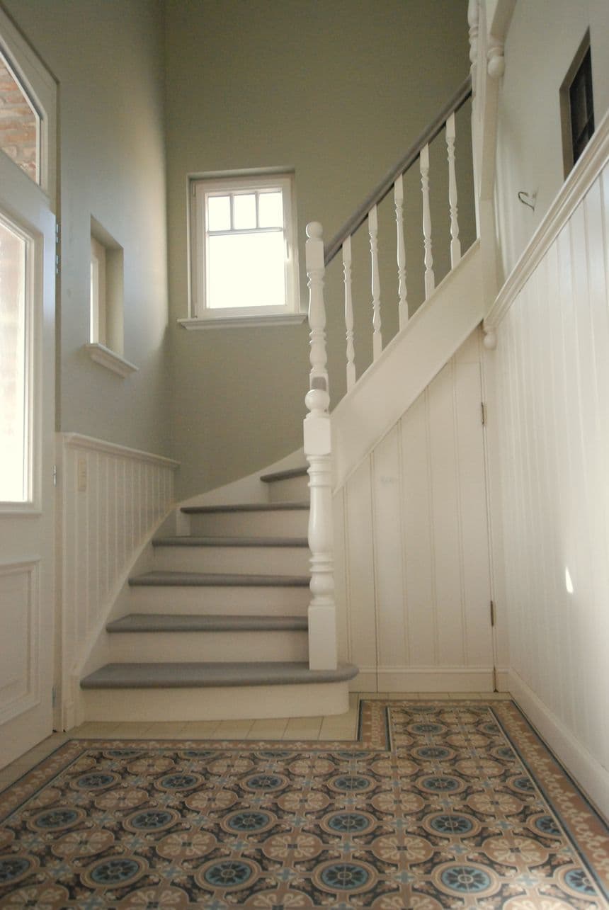 Binnenruimte met witte trap en balustrade, lichtgrijze muren, venster bovenaan en vloer met patroontegels.