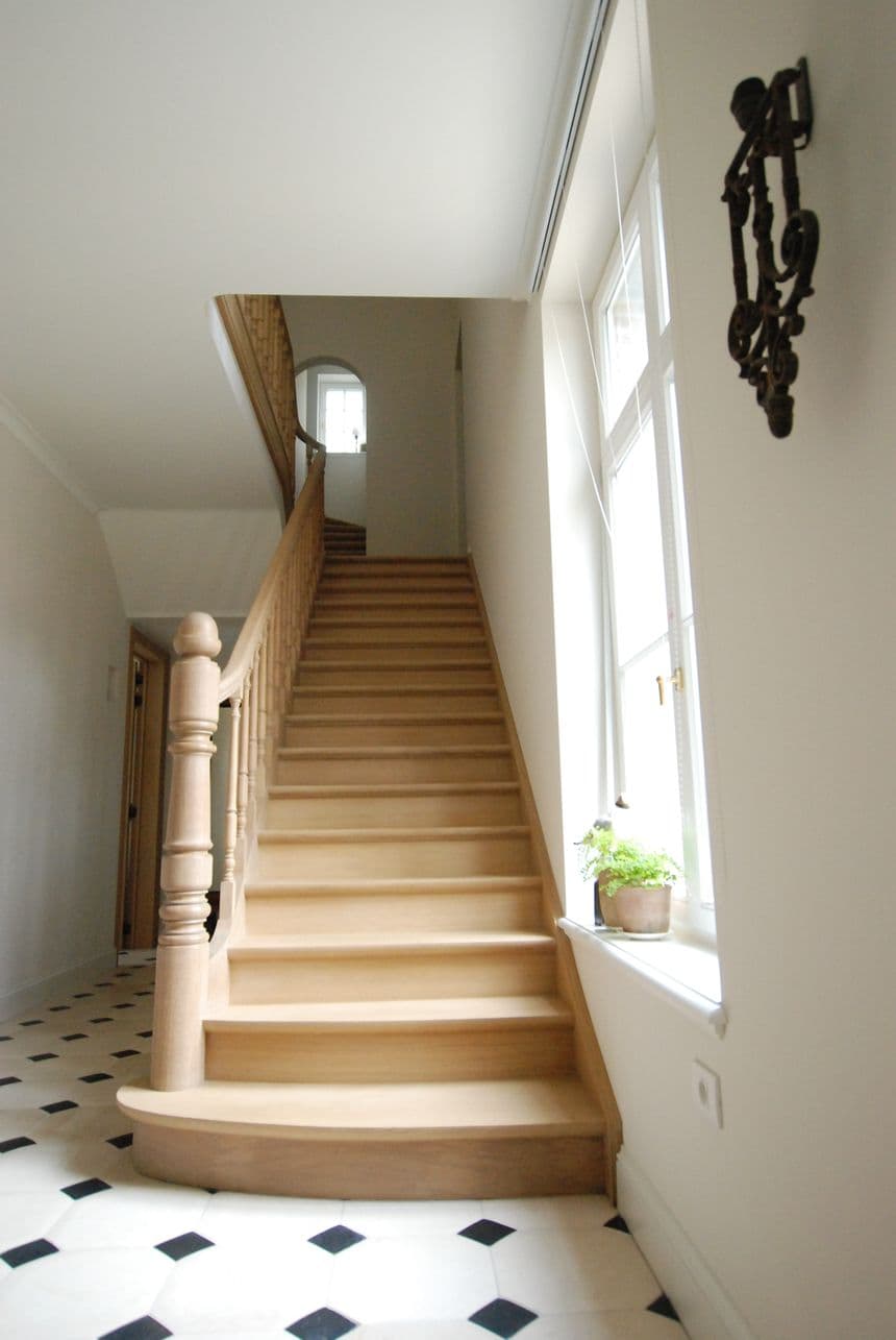 Houten trap met leuning in een lichte hal; venster met plant en een zwart-wit tegelvloer.