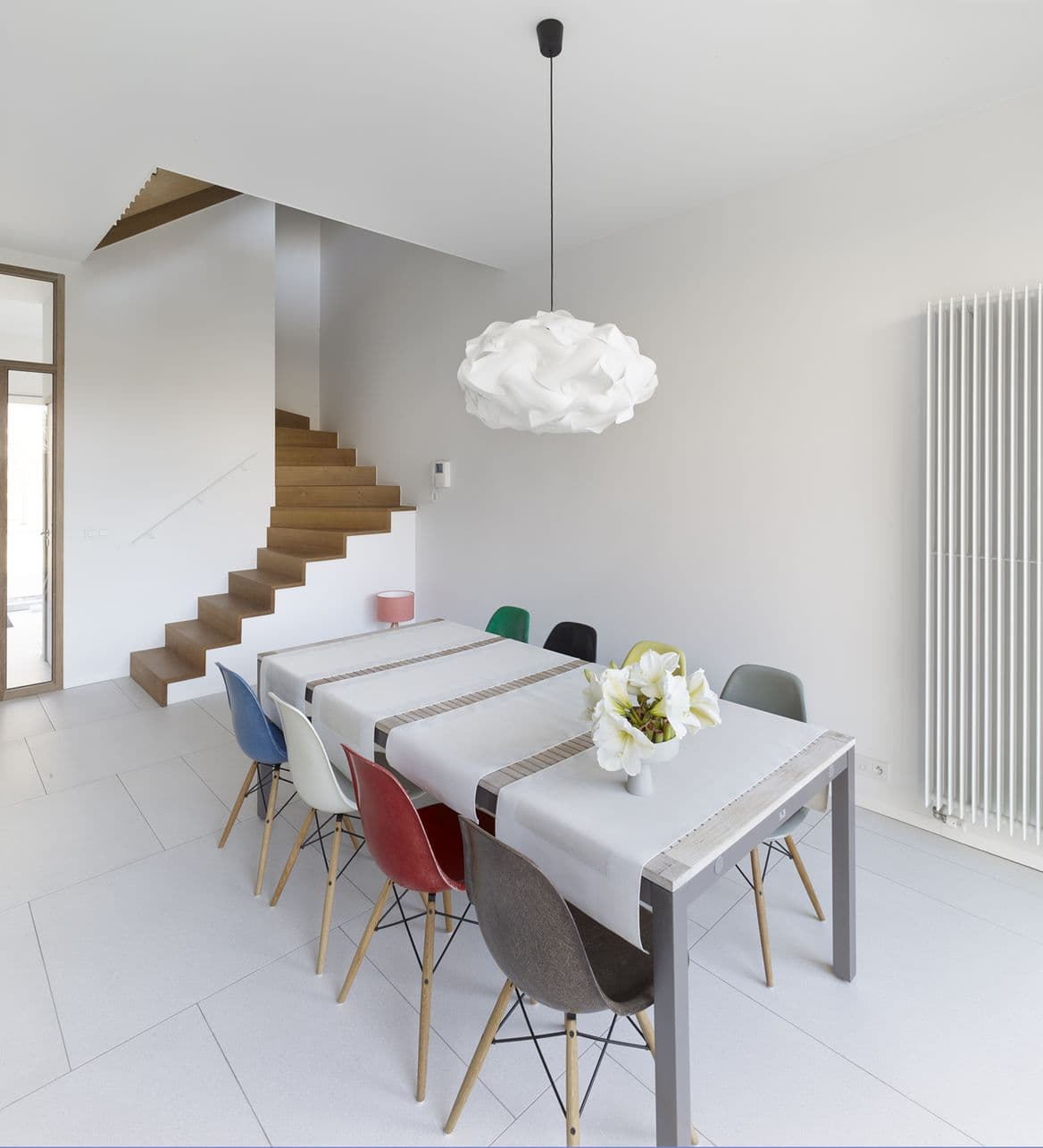 Moderne eetkamer met lange tafel, kleurrijke stoelen, witte wolkenachtige hanglamp, houten trap en vaas met witte bloemen.