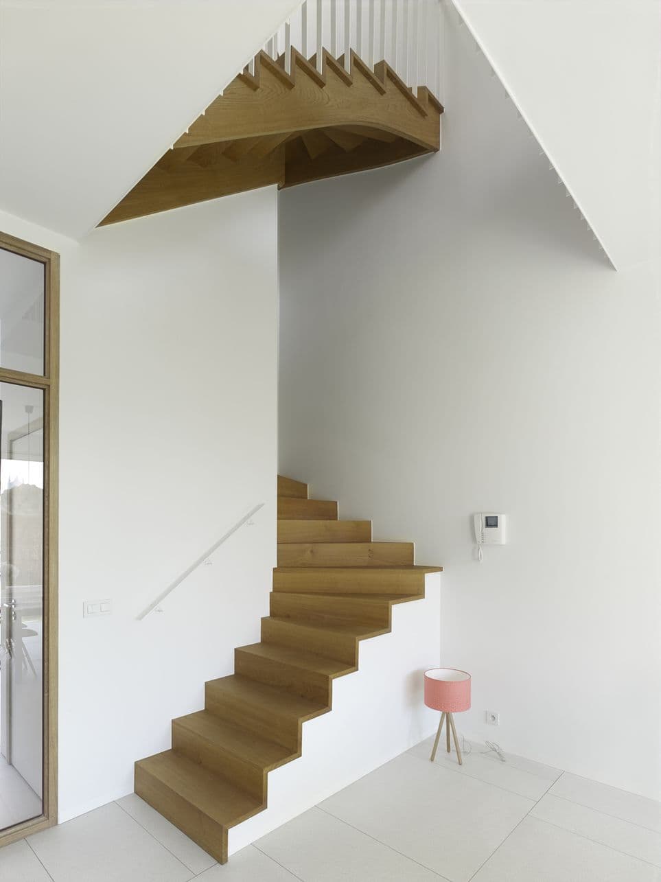 Moderne houten trap met open treden tegen een witte muur; roze lamp op houten pootjes op de vloer.