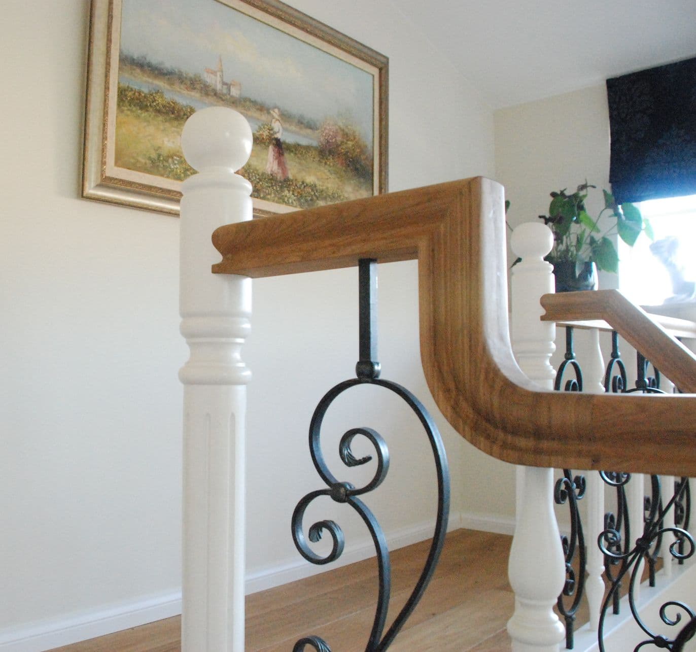 Houten trapleuning met witte newelpalen en zwarte smeedijzeren spijlen, schilderij aan de wand en planten bij het raam.