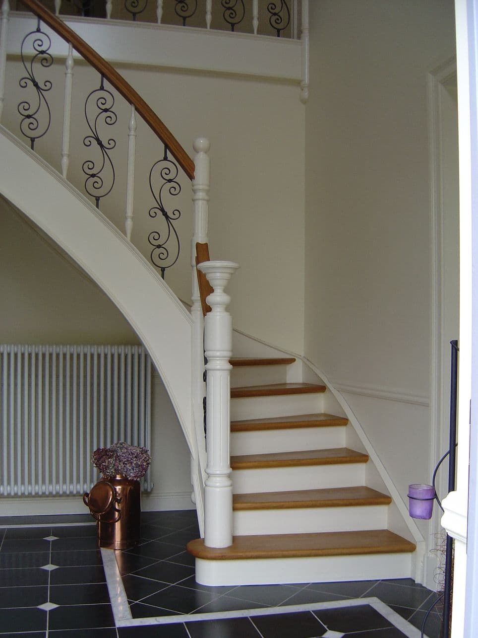 Elegante, witte wenteltrap met houten treden en zwarte smeedijzeren balustrade. Zwart-witte tegelvloer en koperen vaas met droogbloemen links beneden.