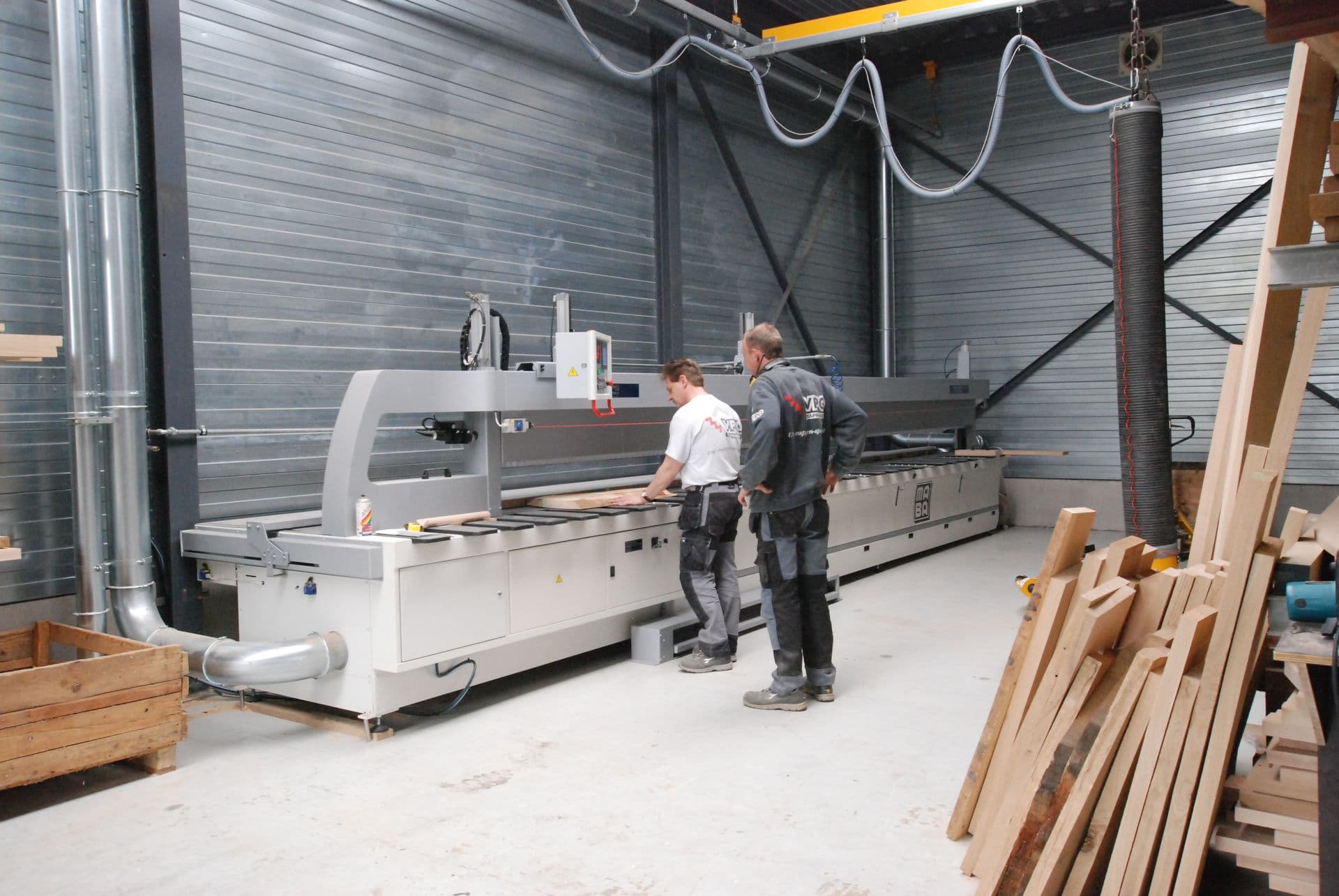 Twee werknemers bedienen een lange houtbewerkingszaagmachine in een atelier, met houten planken en een industriële ruimte.